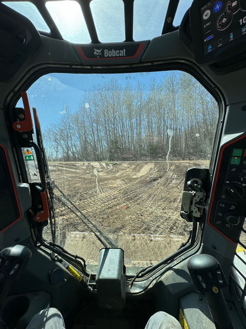 View from inside a Bobcat machinery cab overlooking a cleared worksite and tree line.