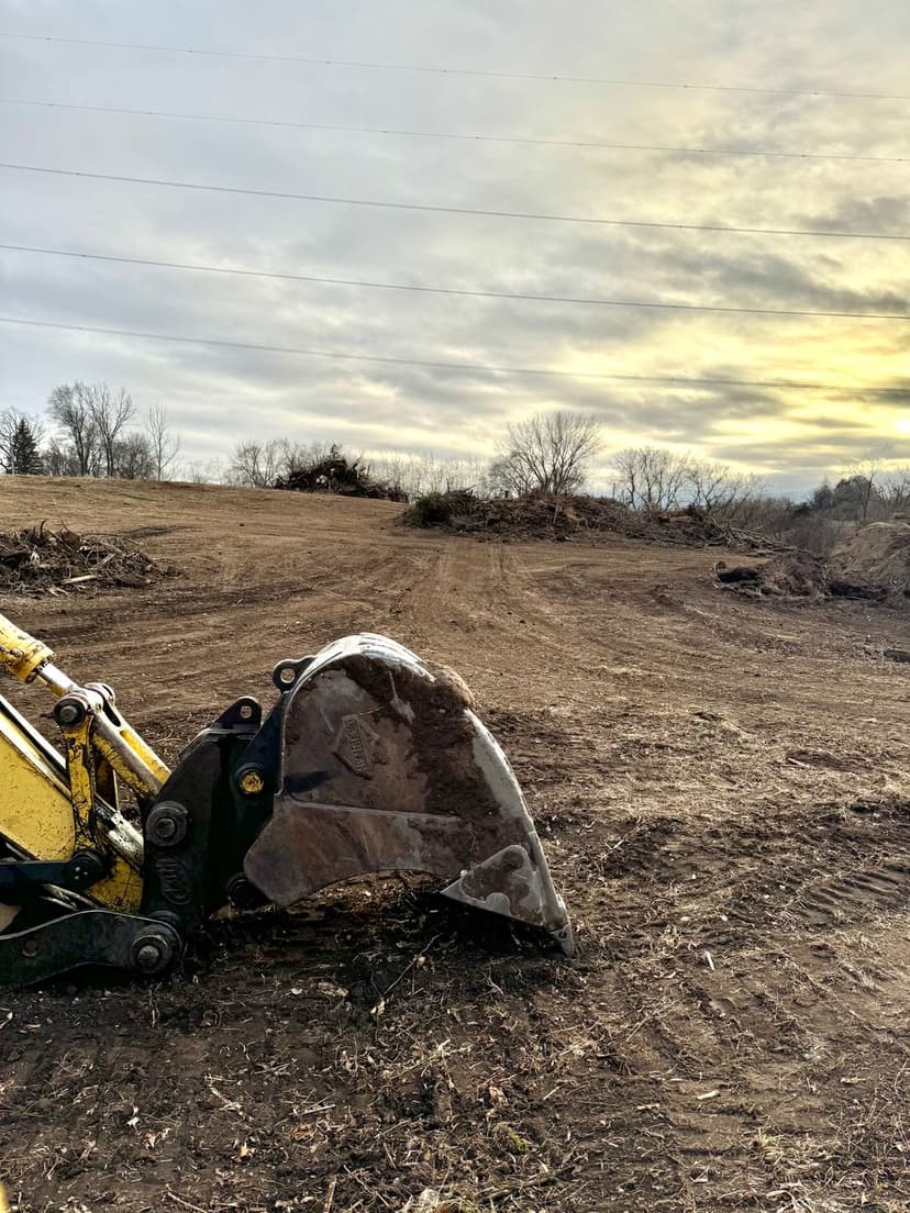 Excavator bucket in a cleared field under a cloudy sky, ready for land development.